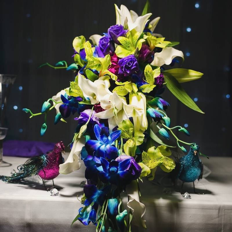 A cascading bouquet of purple roses, white lilies and blue orchids on the head table, with peacock ornaments and twinkle lights