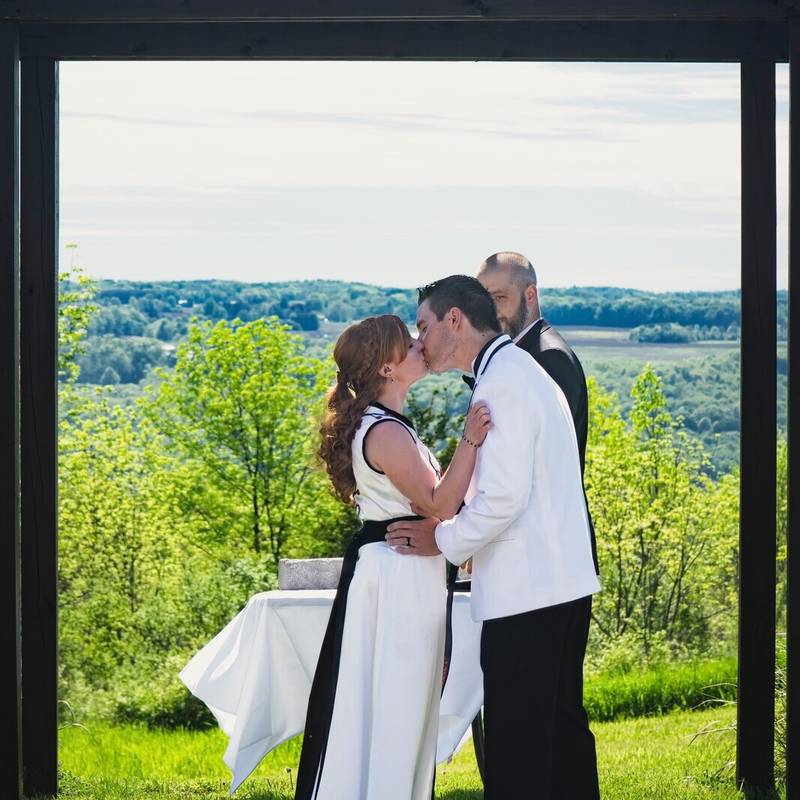 Symphony and Jonathan's ceremony kiss under a dark wooden pergola at the Lakewatch Inn, the officiant behind them and green hills beyond