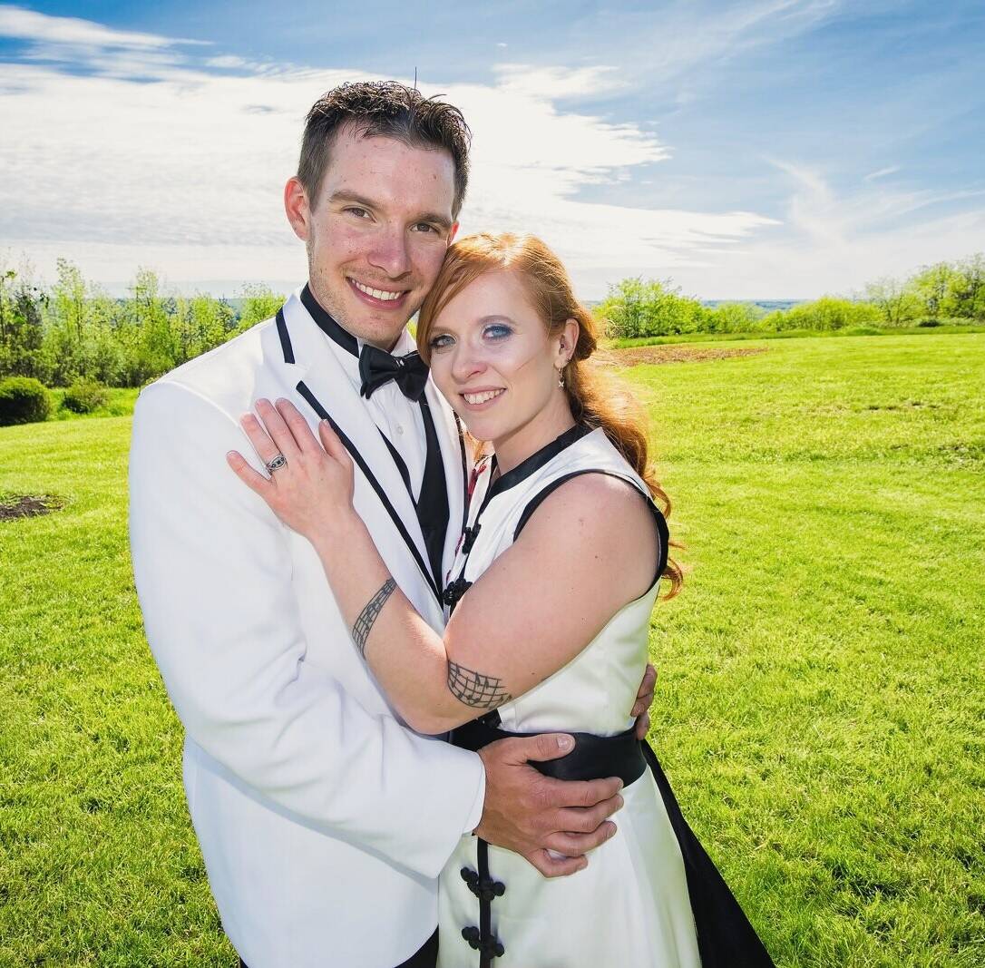 Symphony and Jonathan, in a white tuxedo jacket and a white dress trimmed in black, embrace in a sunny field under a blue sky