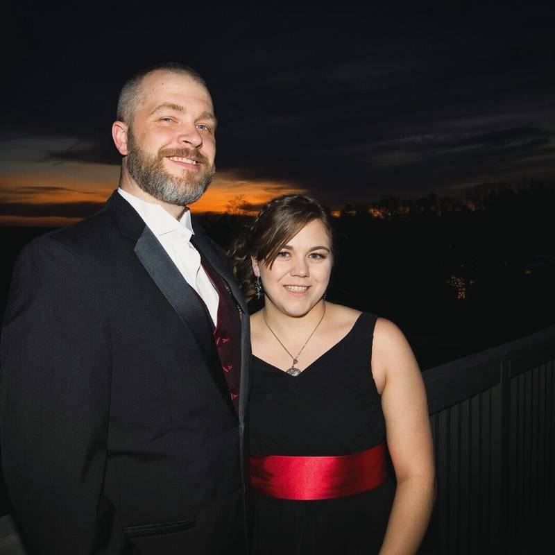 A groomsman in a tuxedo and red vest and a bridesmaid in a black dress with a red sash smile on the deck at sunset