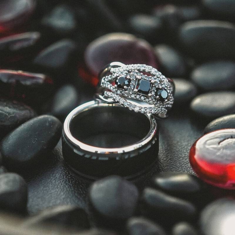 Wedding rings, a silver band set with black and clear stones stacked on a dark band, resting on black pebbles and red glass beads