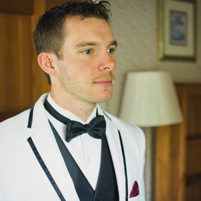 The groom in a white tuxedo jacket with black trim and a black bow tie looks out of frame in a room with a lamp